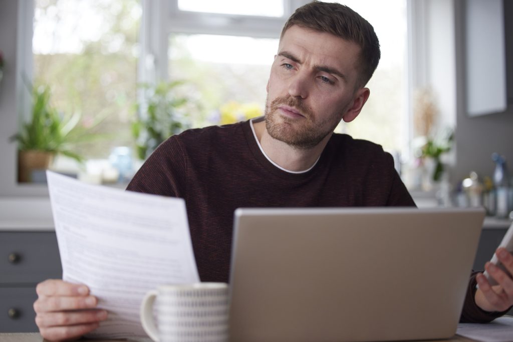 Man In Kitchen At Home Looking At Domestic Bills With Laptop And Calculator